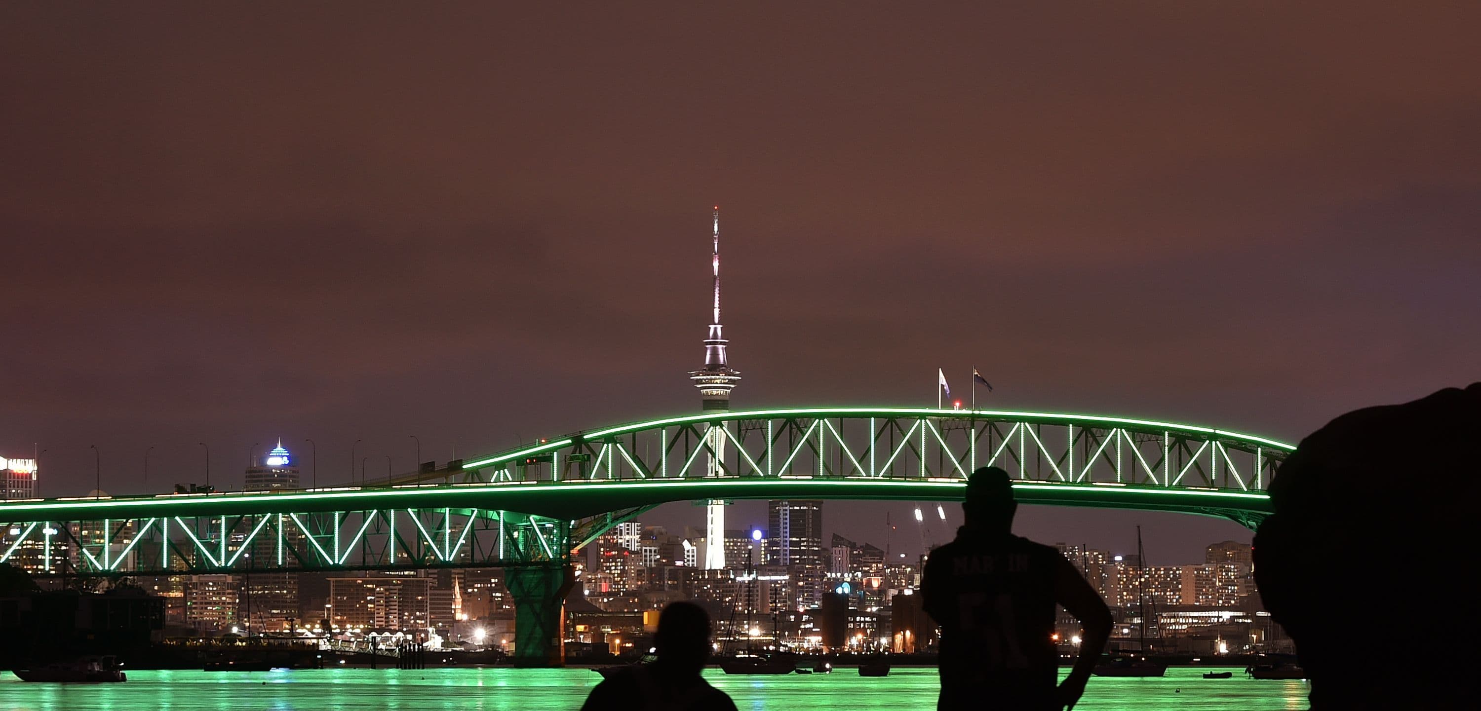 Night view of Auckland harbour bridge lit up in green with silhouette of two people in foreground