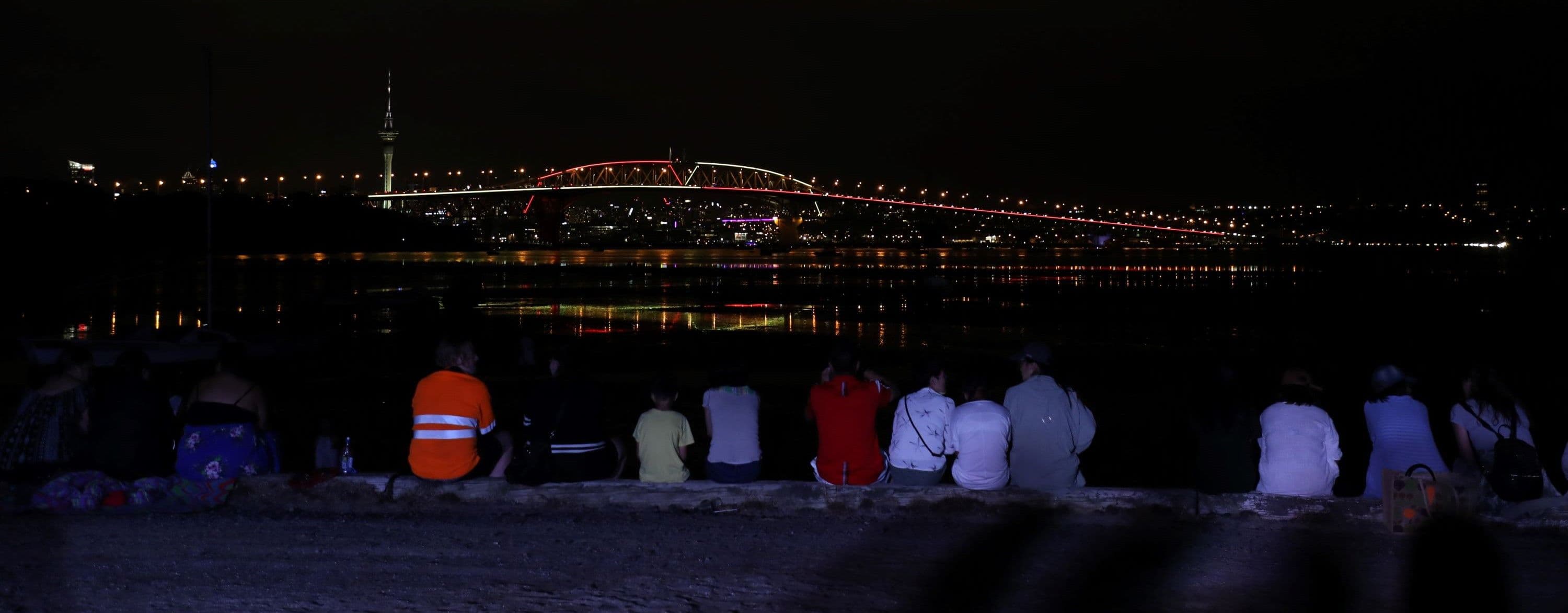 people sitting on wall at night watching harbour bridge light up
